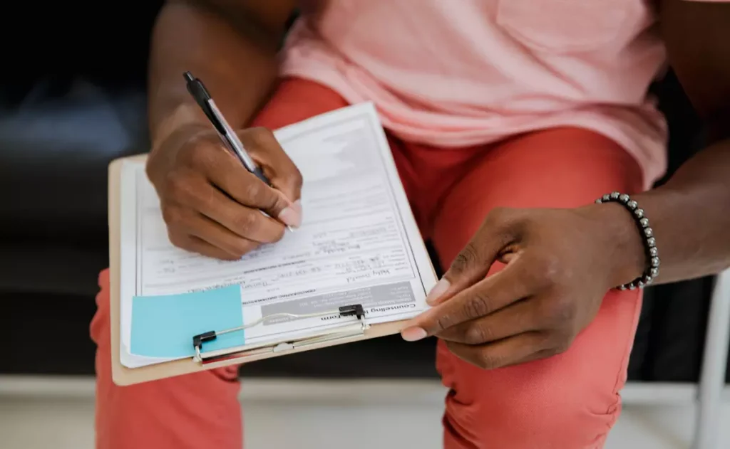 Pessoa sentada preenchendo um formulário em uma prancheta com caneta, segurando o papel com uma das mãos. Na prancheta há um post-it azul preso, e a pessoa usa calça vermelha e camiseta clara.