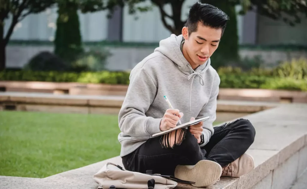 Jovem sentado ao ar livre em um banco de pedra, usando moletom cinza e calça preta rasgada, escreve em um caderno apoiado no colo, com mochila clara ao lado.