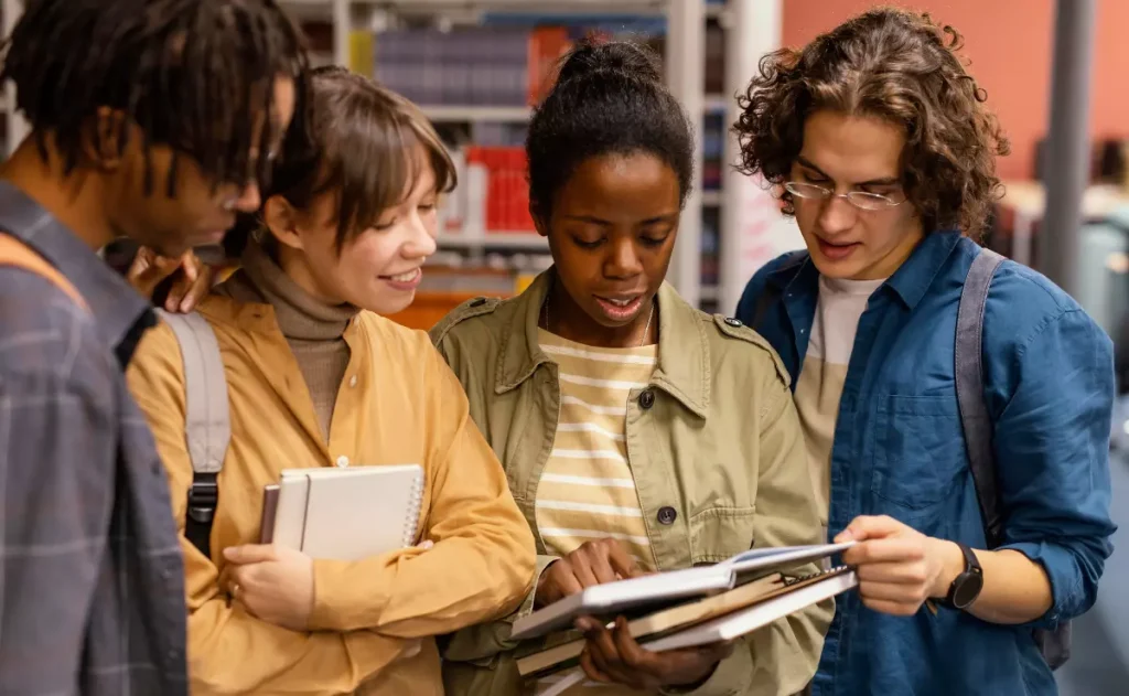 faça alt text dessa imagem Quatro jovens estudantes universitários, de diversas etnias, reunidos em uma biblioteca. Três deles estão olhando para um livro aberto que a quarta estudante, uma jovem negra com cabelo preso, segura e aponta com o dedo.