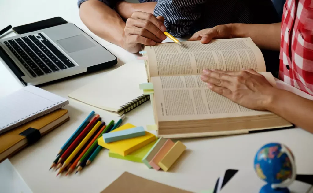 Um estudante com uma caneta amarela aponta para um texto em um dicionário aberto, enquanto outro estudante, sentado ao seu lado com um notebook e materiais escolares como lápis de cor e post-its sobre a mesa, observa.