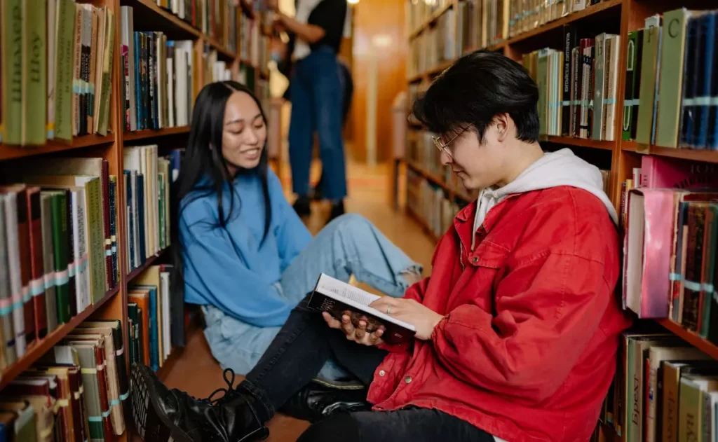 Dois estudantes estão entre as prateleiras de uma biblioteca, enquanto ao fundo, outra estudante se dedica à leitura de livros.