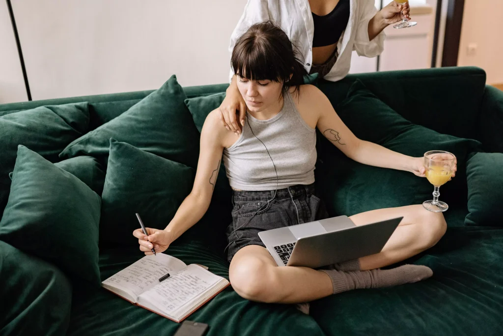 Duas mulheres relaxam no sofá verde, uma com laptop e suco, outra em pé com suco no ombro da amiga.