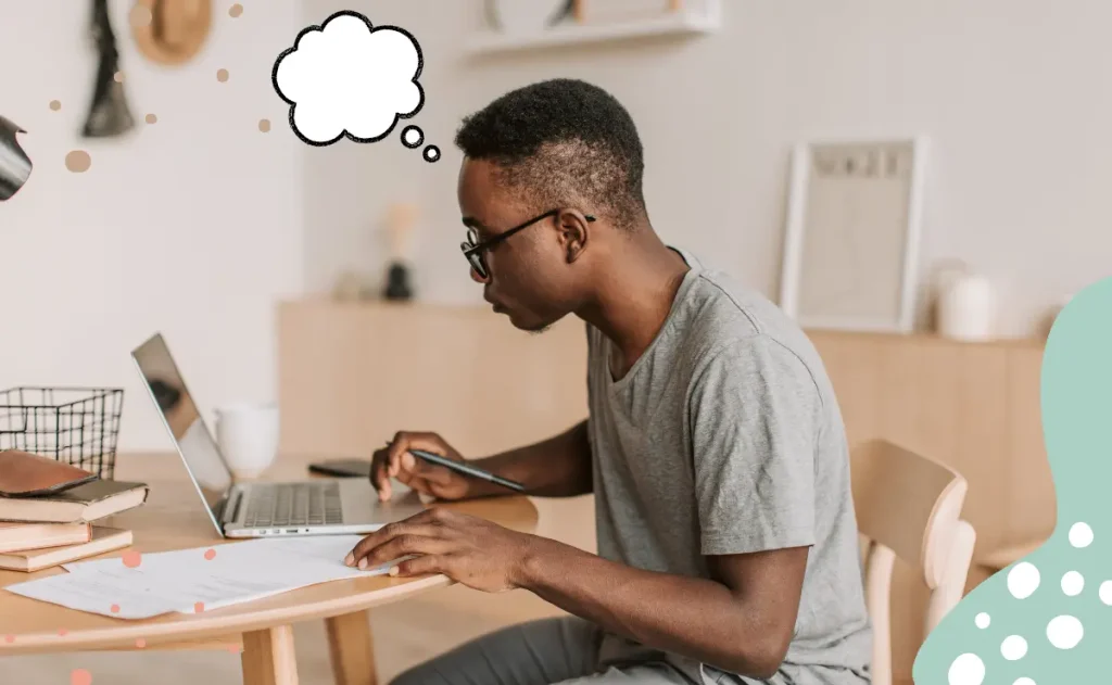 Homem jovem sentado à mesa, usando óculos e camiseta cinza, concentrado em escrever em uma folha de papel enquanto olha para a tela de um notebook. Ao lado, há livros e uma caneca.