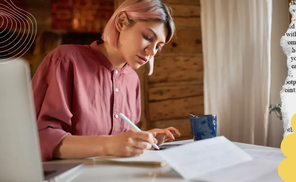 Mulher com cabelo rosa, sentada a uma mesa de madeira, escrevendo num caderno com uma caneta. Há um notebook, uma caneca e alguns papéis na mesa.