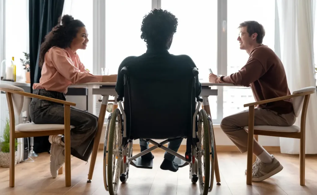 Ao centro, uma pessoa em cadeira de rodas está de costas para o observador, sentada à mesa. Duas outras pessoas a flanqueiam, uma de cada lado, também sentadas à mesa, indicando um ambiente de interação e inclusão.