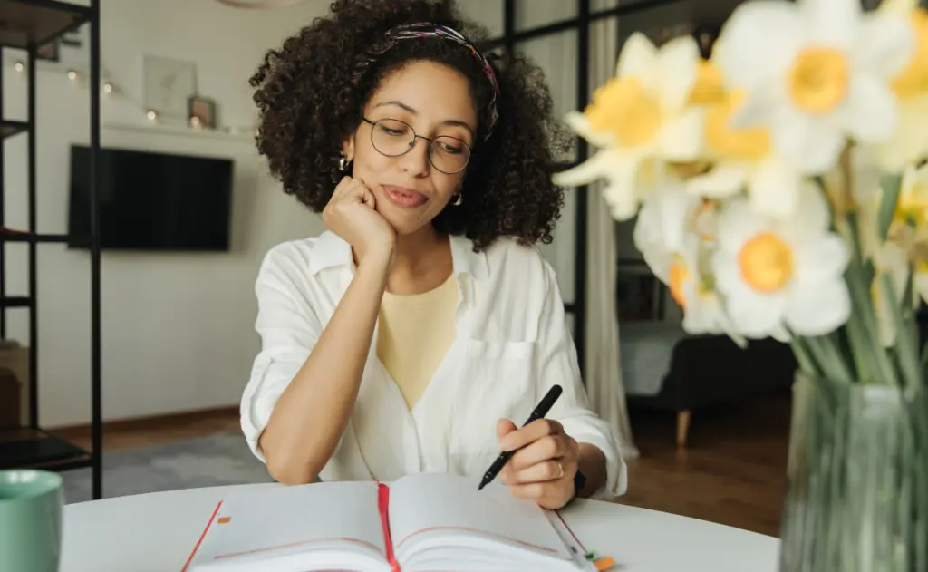 Mulher com cabelo cacheado sentada a uma mesa, usando óculos e uma blusa branca, segurando uma caneta e fazendo anotações em um caderno, com um vaso de flores amarelas desfocado em primeiro plano.