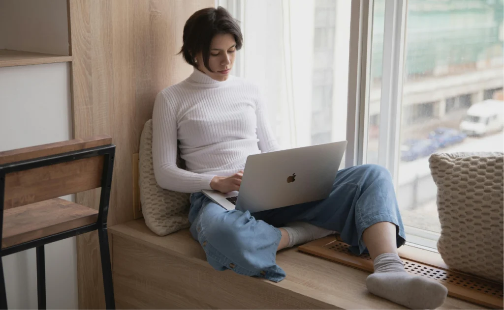 Mulher sentada em uma janela de madeira, vestindo uma blusa de gola alta branca e calças jeans, usando um notebook com a marca da maçã na tampa, com o foco do olhar na tela.