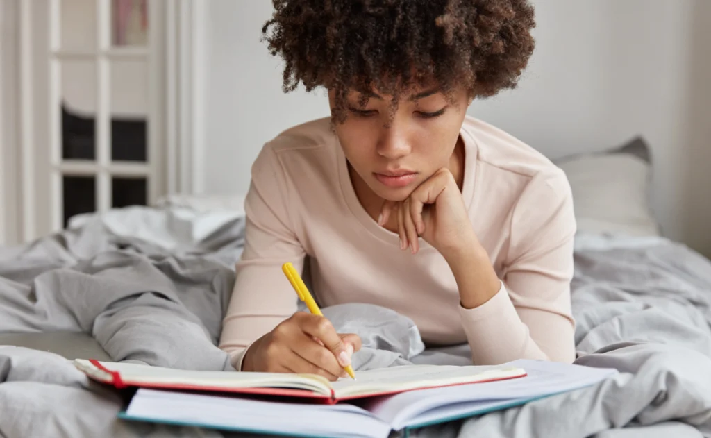 uma mulher de cabelo cacheado deitada em uma cama, com um livro aberto e uma caneta na mão, fazendo anotações.