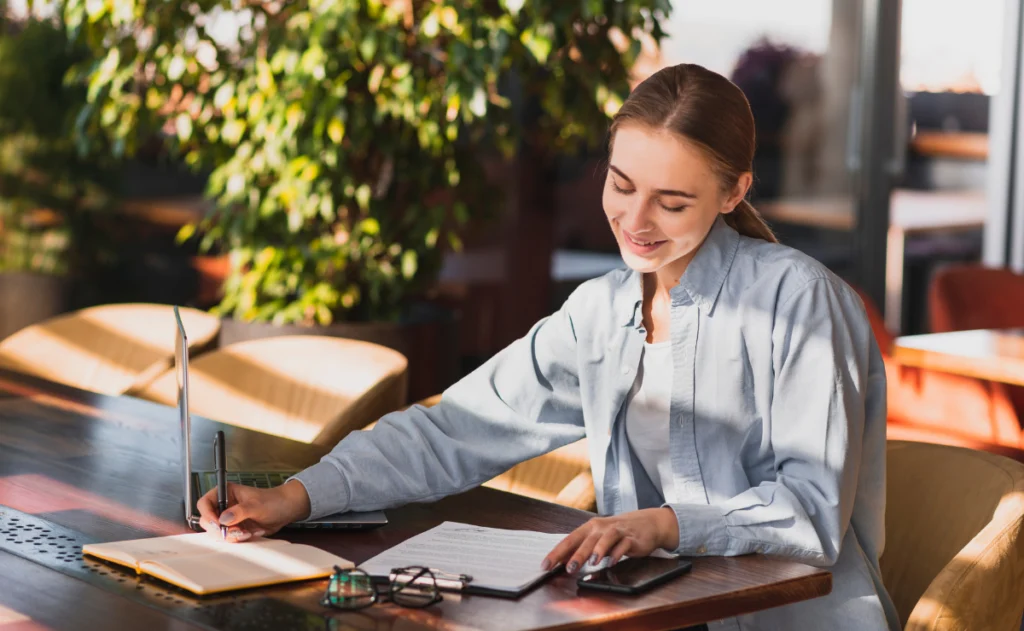 Mulher sorridente de camisa azul escrevendo em um caderno em uma mesa de madeira. A luz do sol da tarde entra pela janela. Na mesa, há um laptop, óculos de grau e um smartphone.