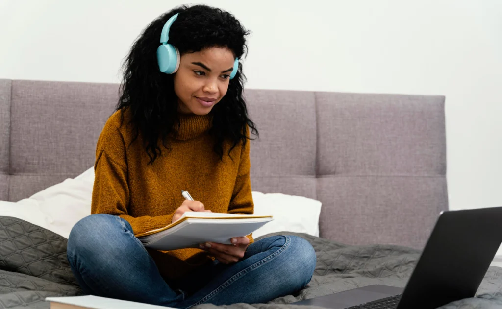 Mulher usa um moletom de malha mostarda e calça jeans. Fones de ouvido azuis claros cobrem suas orelhas. Ela olha para baixo, concentrada, enquanto escreve em um caderno de capa amarela com a mão direita. Em frente a ela, está um notebook preto aberto.