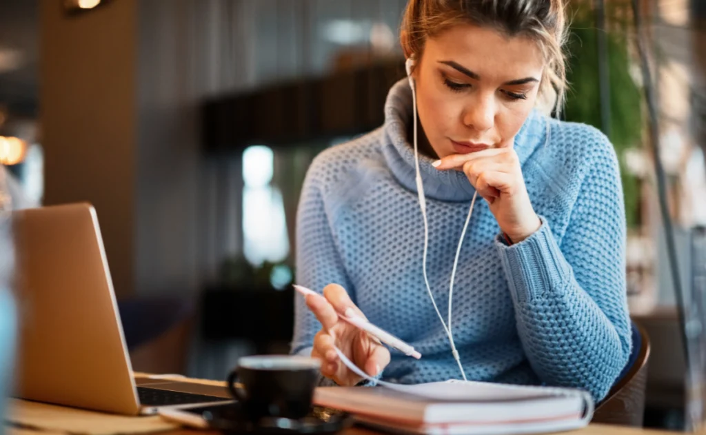 Uma jovem com um suéter azul, fones de ouvido e óculos, está concentrada em seus estudos, com um caderno aberto e um laptop à sua frente.