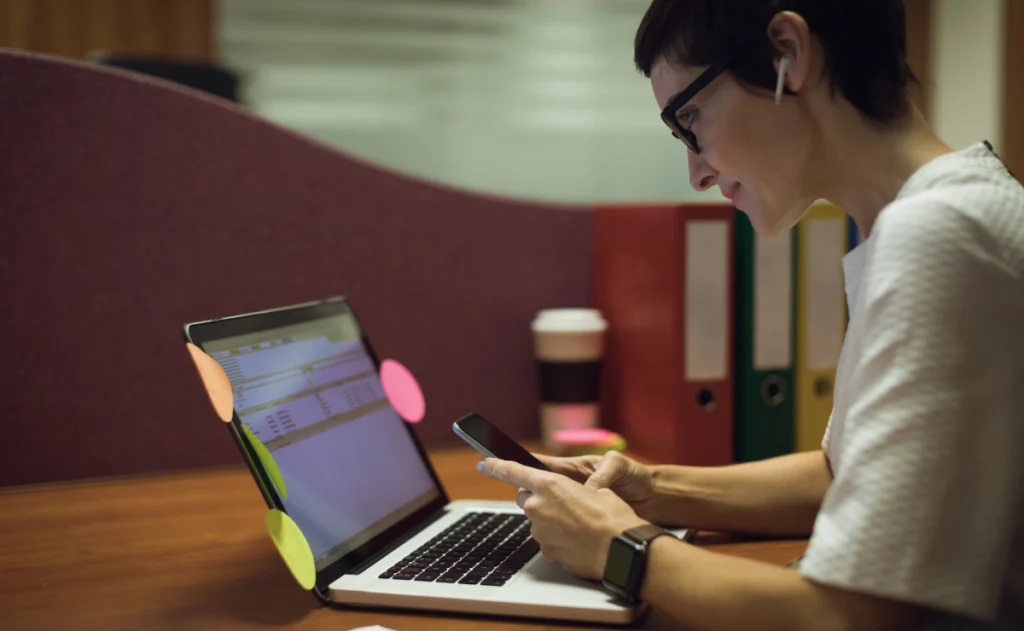 Pessoa sentada em uma mesa de escritório, usando óculos e fones de ouvido sem fio, olhando para o celular enquanto trabalha em um notebook com post-its coloridos colados na tela. Ao fundo há pastas organizadoras e um copo descartável de café.