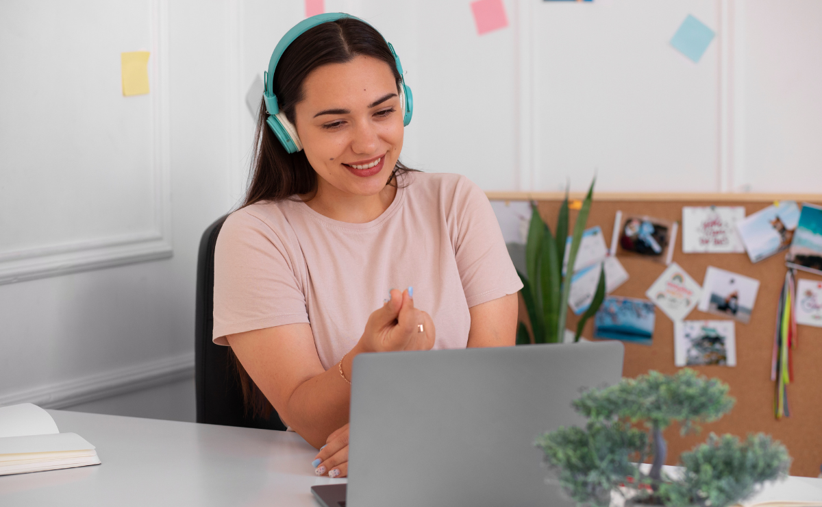 Mulher sorridente com fones de ouvido participa de atividades de escrita e leitura em ensino remoto usando laptop, em ambiente de estudo organizado e acolhedor.