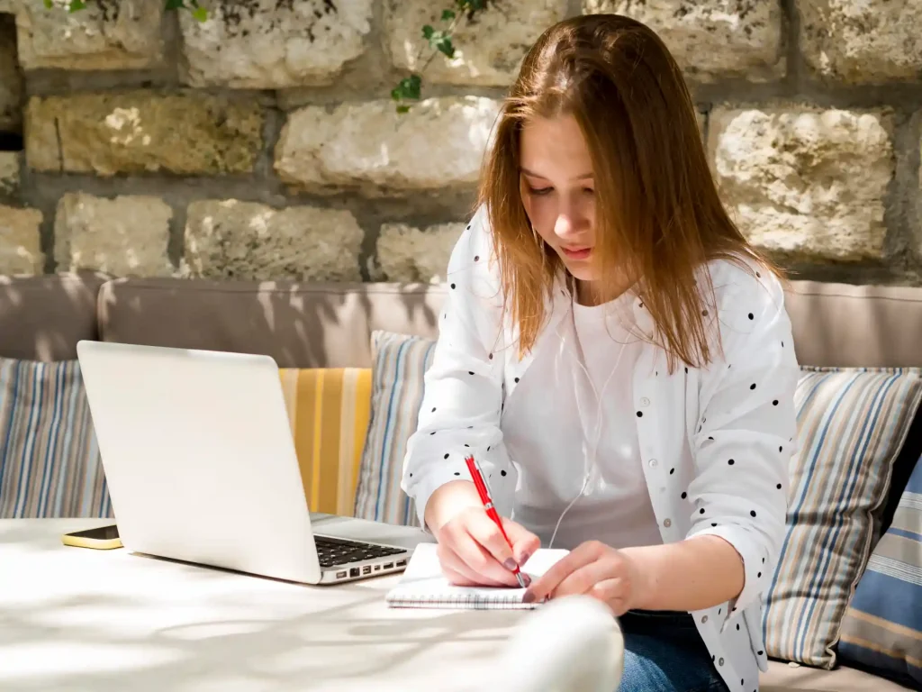 Jovem mulher estudando  em frente a um laptop, escrevendo em caderno com uma caneta vermelha, em ambiente de café ou escritório com parede de pedra ao fundo