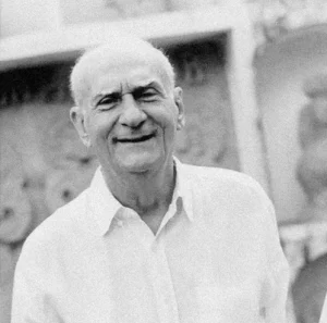 Ariano Suassuna, homem idoso e com poucos cabelos, em foto em preto branco sorrindo usando uma camisa branca