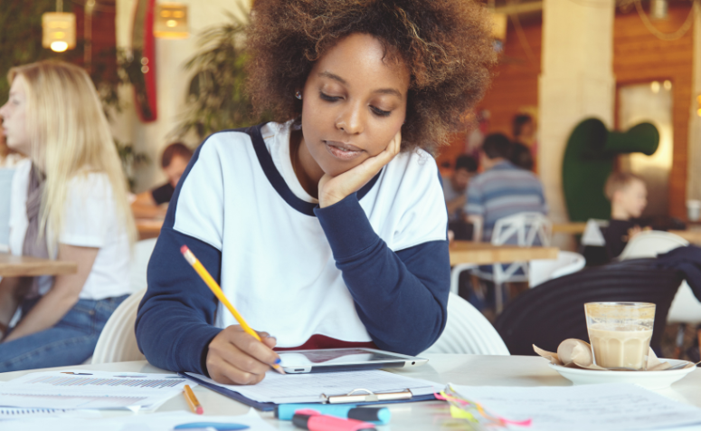 como-usar-repertorio-critico-na-redacao Jovem estudante de aparência pensativa, sentada em uma cafeteria, estudando e fazendo anotações para aprimorar o repertório crítico na redação.