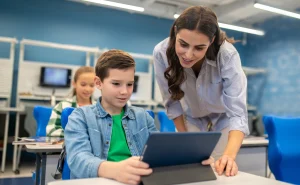 Professora auxiliando estudantes de escola privada com tecnologia na sala de aula, mostrando uso de tablet para aprendizagem interativa.