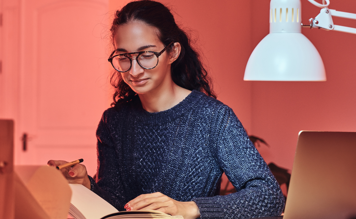 Mulher jovem com óculos e suéter, estudando em uma mesa com laptop e luminária, representando o conceito de repertório sociocultural sobre negacionismo científico.