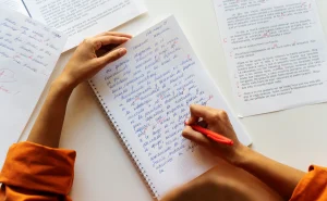 Imagem de uma estudante, em uma mesa branca, focada na mão direita que segura uma caneta vermelha e escreve um texto em um caderno de espiral. A mão esquerda está posicionada na parte superior da folha. O ambiente sugere estudo, talvez para o ENEM ou outro exame.