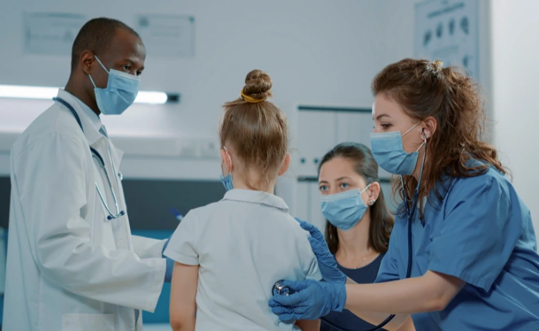 Equipe médica atende uma criança em um consultório. Uma profissional usa estetoscópio para examinar a menina, enquanto outro médico e uma mulher observam.