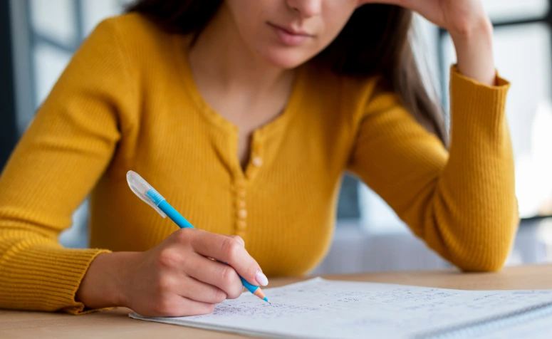 Uma mulher jovem, vestindo uma blusa de tricô amarela, escreve concentradamente em um caderno com uma caneta azul, enquanto apoia a cabeça na outra mão. A imagem foca nas mãos e no caderno sobre uma mesa de madeira clara.