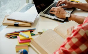 Pessoa estudando em uma mesa com materiais de apoio. Há um laptop aberto, um caderno, lápis coloridos, post-its e um livro sendo lido. A cena mostra um ambiente de estudo organizado e concentrado.