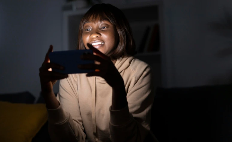 Uma jovem mulher negra, com cabelo curto e franja, sorri enquanto olha atentamente para a tela de um smartphone que segura horizontalmente. A luz forte do aparelho ilumina seu rosto em um ambiente escuro, criando um contraste dramático que sugere o uso noturno ou em baixa luminosidade.