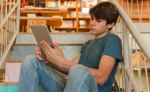 Jovem sentado em uma escada dentro de uma biblioteca, usando uma camiseta azul e calça jeans, enquanto segura um tablet e o observa atentamente. Ao fundo, há estantes com livros, criando um ambiente de estudo e concentração.