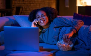Mulher deitada no sofá, sorrindo enquanto fala ao telefone, com um laptop à frente e uma tigela de pipoca ao lado, em um ambiente iluminado em tons de azul e laranja.