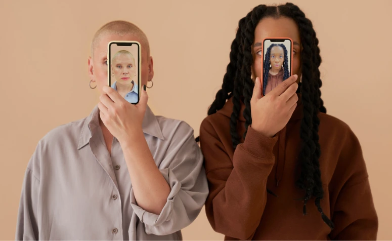 Duas pessoas seguram celulares na frente do rosto, mostrando faces diferentes das delas, representando manipulação e troca de identidades digitais.