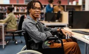Pessoa em cadeira de rodas sorrindo em uma biblioteca, com outras pessoas estudando ao fundo.