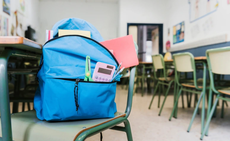 Mochila azul com caderno, lápis, clipes e timer de cozinha sobre uma cadeira em sala de aula, pronta para o dia escolar.